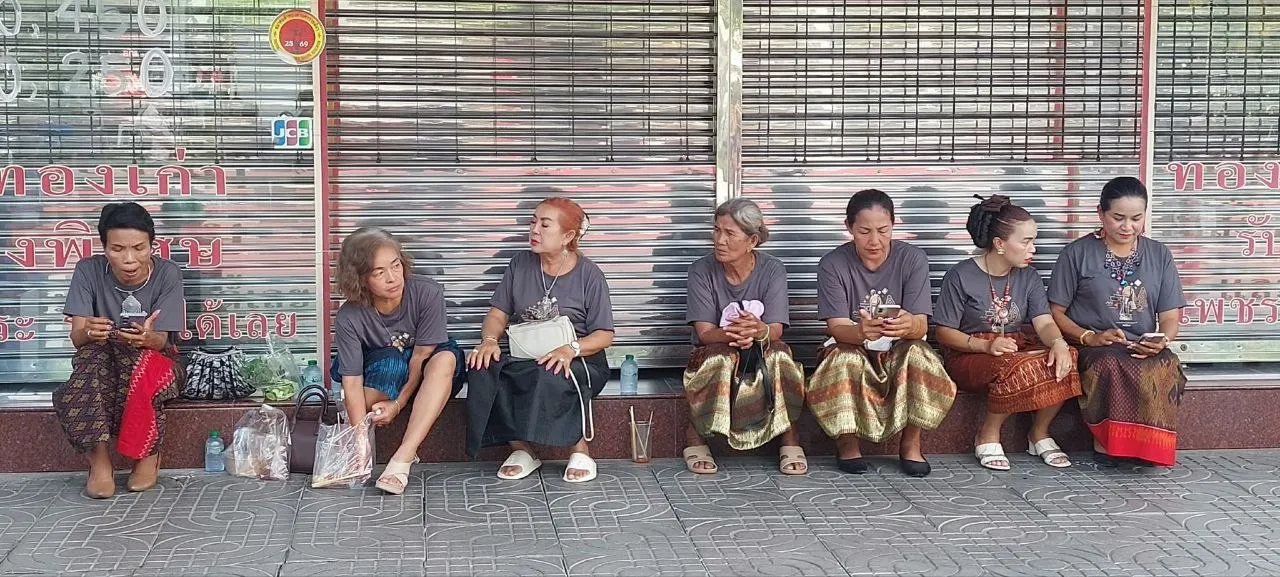 Un gruppo di donne isan con la gonna tradizionale sedute sul marciapiede davanti a un negozio chiuso, tra borse della spesa e cellulari in mano, nel nord-est della Thailandia